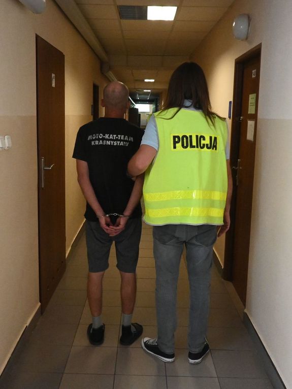 ubrany po cywilnemu policjant w żółtej kamizelce z napisem Policja na plecach prowadzi zatrzymanego korytarzem. Widok z tyłu