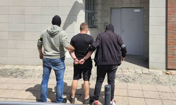 Dwaj ubrani po cywilnemu policjanci stoją z zatrzymanym mężczyzną przed aresztem śledczym. Widok z tyłu