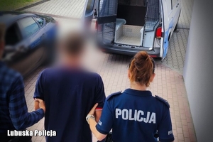 zdjęcie przedstawia dwóch funkcjonariuszy prowadzących mężczyznę z założonymi kajdankami na ręce trzymane z tyłu,  w kierunku otwartych drzwi oznakowanego radiowozu