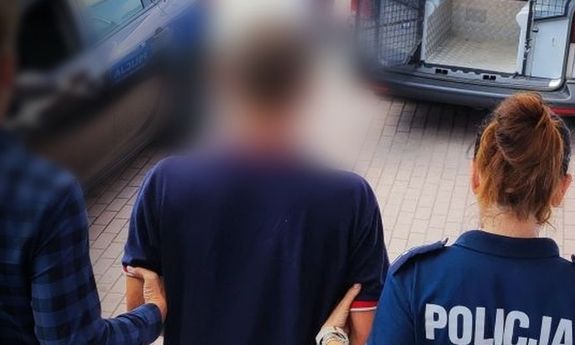 zdjęcie przedstawia dwóch funkcjonariuszy prowadzących mężczyznę z założonymi kajdankami na ręce trzymane z tyłu,  w kierunku otwartych drzwi oznakowanego radiowozu