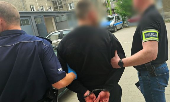 dwaj policjanci, jeden w mundurze, drugi w ubraniu cywilnym, prowadzą zatrzymanego