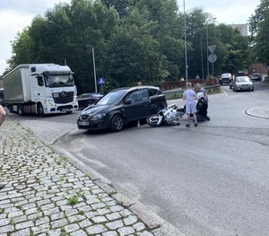 miejsce wypadku. Na drodze przy czarnym samochodzie leży porzewrócony motocykl. W głebi samochody na drodze