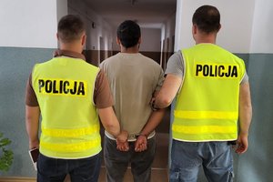 postacie ustawione tyłem do fotografującego, zatrzymany mężczyzna skuty kajdankami z tyłu, pomiędzy dwoma policjantami po cywilnemu w kamizelach z napisem Policja na plecach