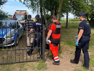 po lewej stronie widać policyjny radiowóz, za nim ambulans przy którym stoi ratownik medyczny. W jego stronę przez furtkę wychodzi mężczyzna, za nim policjantka, ratownik medyczny i policjant