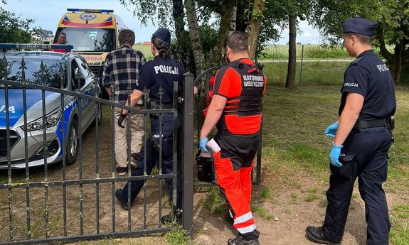 po lewej stronie widać policyjny radiowóz, za nim ambulans przy którym stoi ratownik medyczny. W jego stronę przez furtkę wychodzi mężczyzna, za nim policjantka, ratownik medyczny i policjant