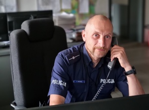 mł. asp. Łukasz Wróbel w mundurze przy biurku, przy uchu ma słuchawkę telefonu stacjonarnego