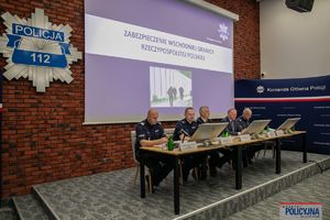 na pierwszym planie komendant i 2 ministrów siedzą przy stole, w tle leci prezentacja