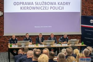 na pierwszym planie ludzie słuchający odprawy, na drugim komendant i 2 ministrów siedzą przy stole, w tle leci prezentacja