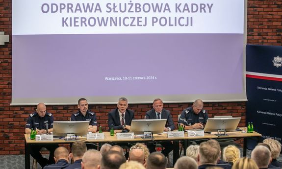 na pierwszym planie ludzie słuchający odprawy, na drugim komendant i 2 ministrów siedzą przy stole, w tle leci prezentacja
