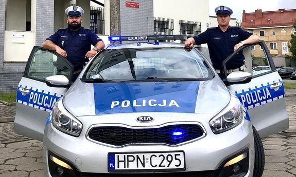 dwaj umundurowani policjanci stoją w otwartych drzwiach radiowozu, po jego lewej i prawej stronie