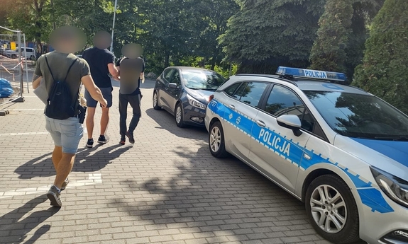 dwóch nieumundurowanych policjantów idzie z zatrzymanym, mającym założone kajdanki,do radiowozu