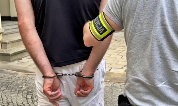 widziany od tyłu zatrzymany mężczyzna zakuty w kajdanki jest prowadzony przez nieumundurowanego policjanta
