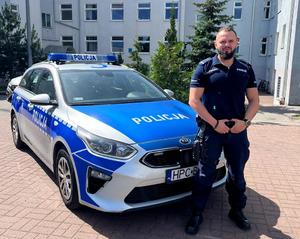 Policjant na tle budynku komendy i radiowozu oznakowanego
