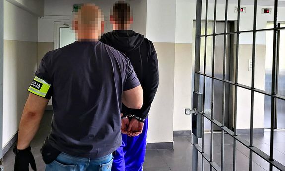 policjant prowadzi zatrzymanego mężczyznę z założonymi na ręce kajdankami