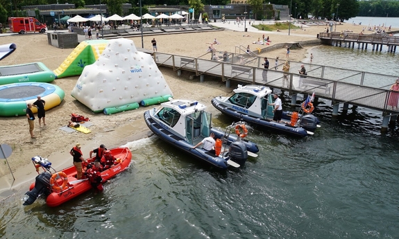 dwie łodzie policyjne i łódź ratowników przy Plaży Miejskiej nad Jeziorem Ukiel, w głębi ludzie wypoczywający na plaży