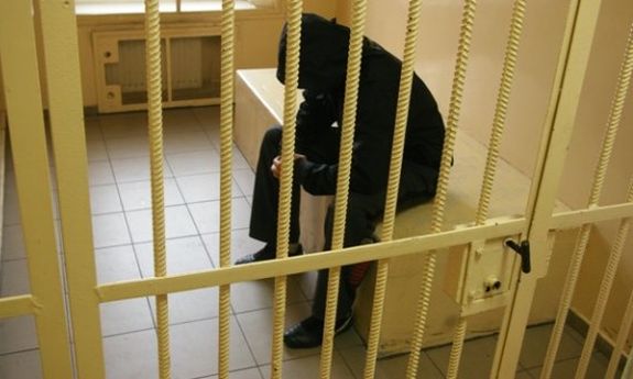 mężczyzna siedzący w celi  ubrany na ciemno z kapturem na głowie
