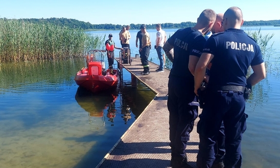 na zdjęciu umundurowani policjanci i strażacy stoją na pomoście, obok pomostu widać łódkę wykorzystywaną do poszukiwań zaginionej kobiety