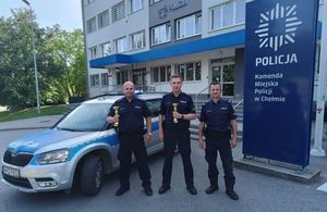 trzej policjanci umundurowani przed budynkiem komendy przy radiowozie, dwóch z nich trzyma w ręku puchary