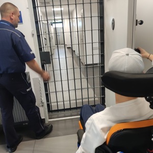 chłopiec na wózku przed pomieszczeniem dla osób zatrzymanych. Kratę otwiera mu umundurowany policjant