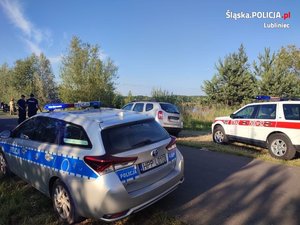 Na zdjęciu widać oznakowany policyjny radiowóz oraz samochód straży pożarnej, w tle zbiornik wodny