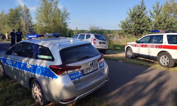 Na zdjęciu widać oznakowany policyjny radiowóz oraz samochód straży pożarnej, w tle zbiornik wodny