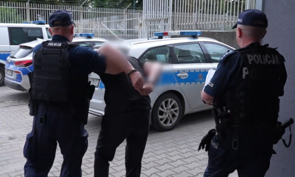 dwóch umundurowanych policjantów prowadzi zatrzymanego mężczyznę do radiowozu