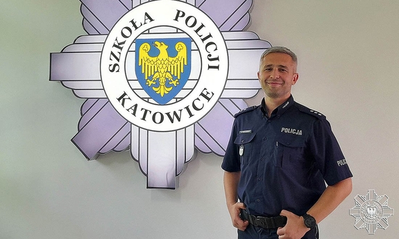 Komisarz Krystian Wieczorek stoi na tle loga Szkoły Policji w Katowicach wiszącego na białej ścianie