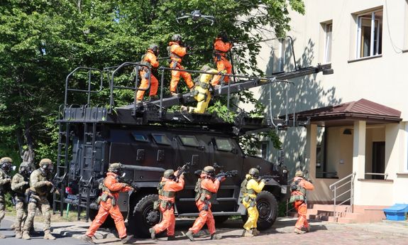 policjanci w specjalnych skafandrach idą do budynku mieszkalnego, obok stoi opancerzony wóz policyjny na którym stoją inni policjanci w specjalnych skafandrach o kolorze pomarańczowym
