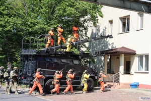 policjanci w specjalnych skafandrach idą do budynku mieszkalnego, obok stoi opancerzony wóz policyjny na którym stoją inni policjanci w specjalnych skafandrach o kolorze pomarańczowym