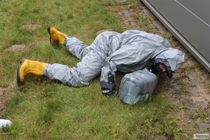 na ziemi leży mężczyzna w białym stroju i żółtych kaloszach z plastikową 5 litrową butelką