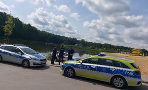 Dwa radiowozy stojące obok akwenu wodnego za nimi trójka, odwróconych plecami policjantów.