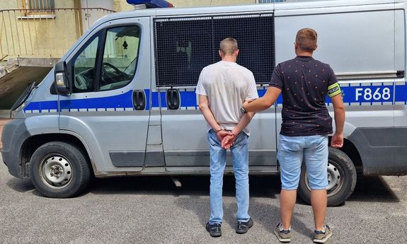 zatrzymany mężczyzna stojący tyłem trzymany za ramię przez nieumundurowanego policjanta na tle radiowozu