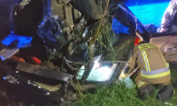 na zdjęciu miejsce wypadku drogowego, obok pojazdu w rowie klęczy strażak i udziela pomocy poszkodowanemu w wypadku