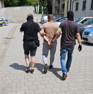 policjanci ubrani po cywilnemu prowadzą mężczyznę z założonymi kajdankami na ręce trzymane z tyłu