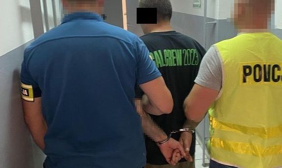 Dwaj nieumundurowani policjanci prowadzą zatrzymanego. Jeden z funkcjonariuszy ma na sobie żółtą kamizelkę z napisem: Policja