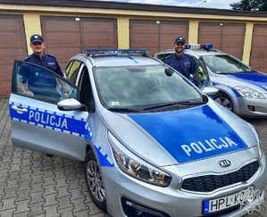 dwóch umundurowanych funkcjonariuszy stoi przy radiowozie