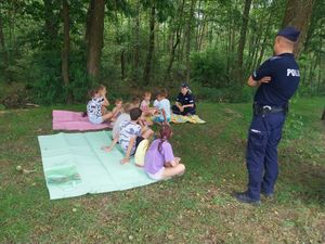 Para policjantów rozmawia z dziećmi siedzącymi na kocach rozłożonych na polanie.