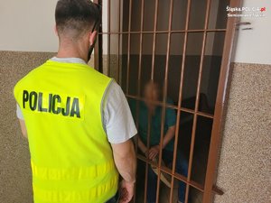 policjant w żółtej kamizelce z napisem: Policja na plecach, stoi przy celi, w której jest zatrzymany