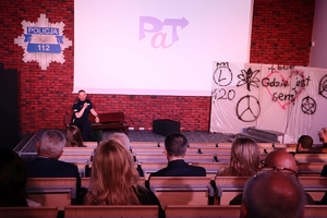 Komendant Główny Policji przemawia do zgromadzonych na widowni gości siedząc na ławce ustawionej na scenie