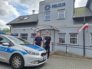 dwaj policjanci stoją przy radiowozie policyjnym zaparkowanym przy budynku komendy policji