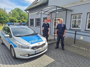 dwaj policjanci stoją przy radiowozie policyjnym zaparkowanym przy budynku komendy policji