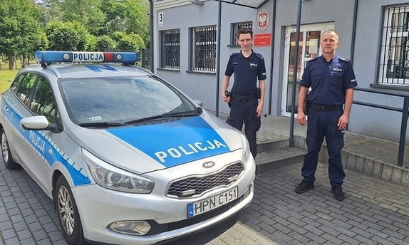 dwaj policjanci stoją przy radiowozie policyjnym zaparkowanym przy budynku komendy policji