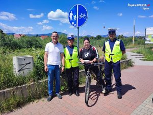 policjanci w umundurowaniu i w odblaskowych kamizelkach , rowerzystka, mężczyzna w białej koszulce a w tle znak drogowy informujący o drodze pieszo-rowerowej