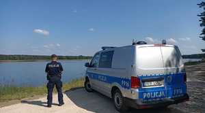 Policjant obrócony w stronę zbiornika wodnego obok stoi radiowóz.