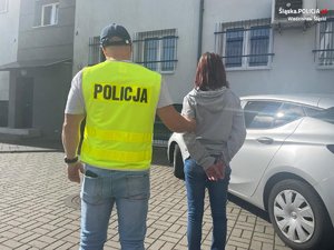 na zdjęciu policjant w odblaskowej kamizelce prowadzi do budynku zatrzymaną kobietę w kajdankach