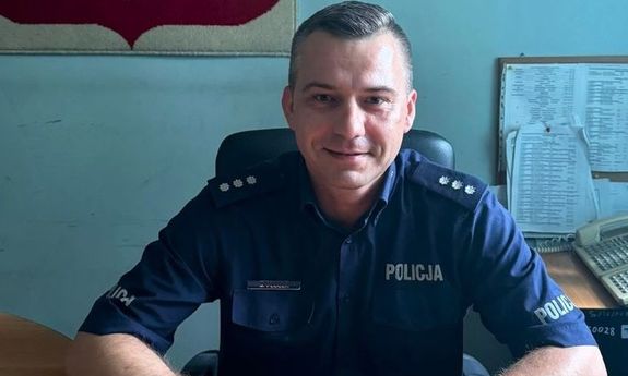 zdjęcie policjanta w umundurowaniu służbowym siedzącego za biurkiem w pomieszczeniu służbowym