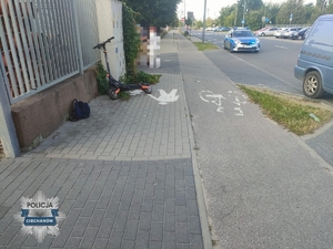 hulajnoga oparta o ogrodzenie przy ulicznym chodniku, na ulicy radiowóz