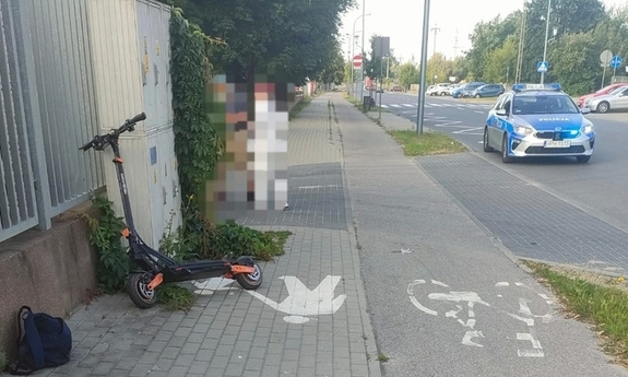 hulajnoga oparta o ogrodzenie przy ulicznym chodniku, na drodze radiowóz