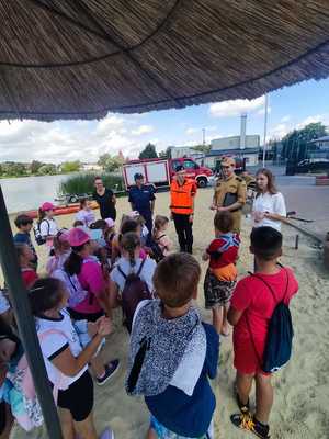 Policjantki, strażak i pracownica Sanepid-u prowadzą na plaży prelekcję dla grupy dzieci.