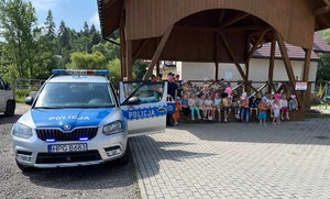 Policjant pozuje do zdjęcia z grupą dzieci obok po lewej stronie stoi radiowóz.